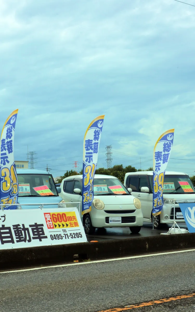 中古車が並ぶ店舗前で、青いのぼりが風に揺れる販売店の風景